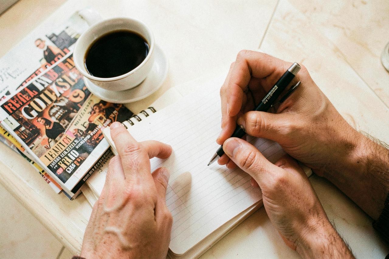 Hands writing in a notebook beside coffee and stacked culture magazines